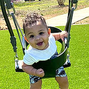 Amarion is registered to the contest to win money with this photo: baby, child, swing, playground, outdoor, grass, smiling, happy, curly_hair, white_shirt, shorts, sunny, park, seat, metal_chains, cute, infant, person, summer, fun