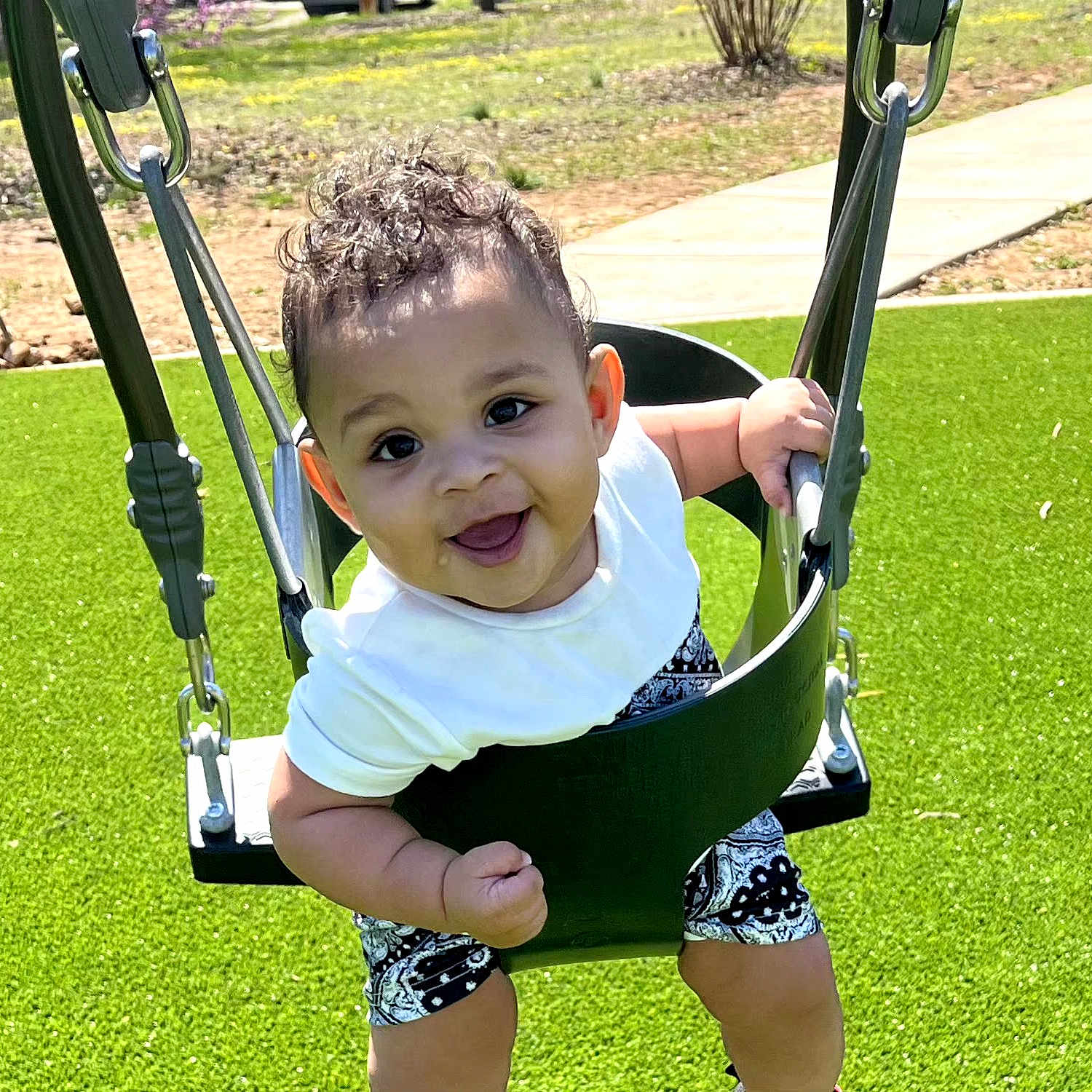 Amarion is registered to the contest to win money with this photo: baby, child, swing, playground, outdoor, grass, smiling, happy, curly_hair, white_shirt, shorts, sunny, park, seat, metal_chains, cute, infant, person, summer, fun