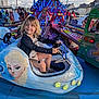 amusement_park, blonde_hair, blue, building, car, carnival_ride, child, clothing, daytime, entertainment, frozen_character, fun, happy, lights, outdoor, people, play, seat, smiling, steering_wheel
