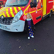 Lëila participe au concours pour gagner de l'argent avec cette photo : black_shoes, child, costume, daylight, door, emergency_vehicle, headlight, license_plate, long_hair, outdoor, parking_area, pavement, person, red_vehicle, side_mirror, smiling, toy, vehicle, window, yellow_stripes