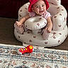 baby, red_hat, inflatable_chair, teddy_bear_pattern, smiling, toy_microphone, carpet, indoor, child, cute, happy, seated, playful, infant, footwear, pink_clothing, person, young_child, soft_toys, home