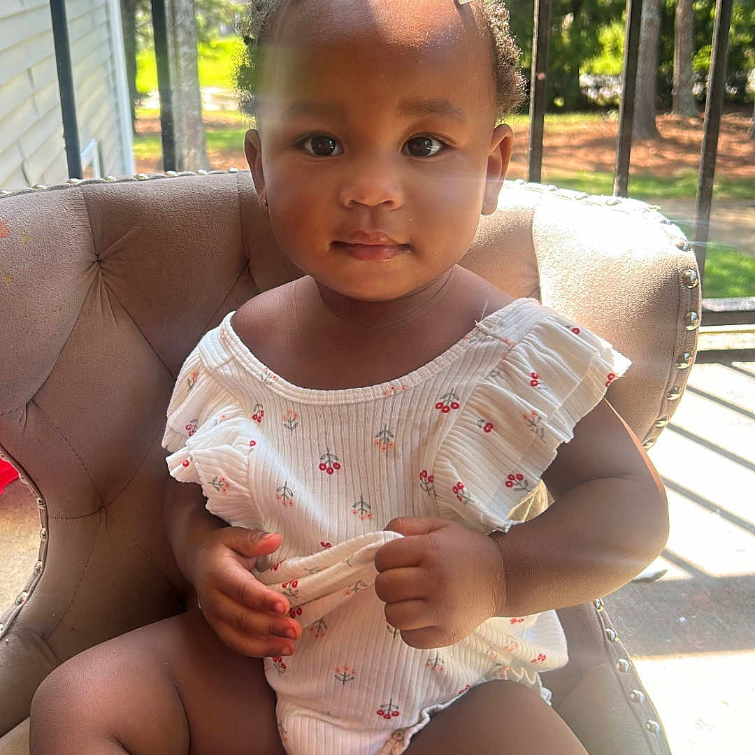 Ariyah is registered to the contest to win money with this photo: toddler, child, baby, portrait, sitting, chair, outdoor, porch, sunlight, trees, balcony, railing, dress, romper, bow, smiling, face, hands, legs, family_photo