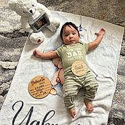 Yahvonta is registered to the contest to win money with this photo: baby, blanket, teddy_bear, wooden_plaques, infant, person, clothing, striped_pants, floor_rug, cute, portrait, baby_milestone, lying_down, home_interior, soft_toy, baby_boy, script_text, face, newborn, child