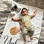baby, blanket, teddy_bear, wooden_plaques, infant, person, clothing, striped_pants, floor_rug, cute, portrait, baby_milestone, lying_down, home_interior, soft_toy, baby_boy, script_text, face, newborn, child