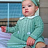 baby, child, knitted_clothing, beret, green_outfit, sitting, blanket, window, indoor, portrait, cute, face, earring, soft_toys, expression, person, footwear, hand, furniture, cozy