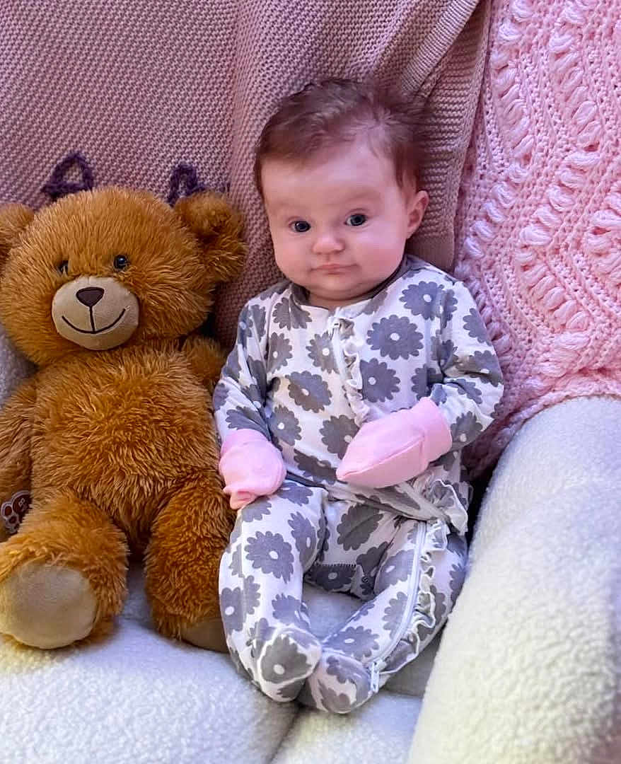 Amelia joined the competition — help win amazing prizes! baby, teddy_bear, blanket, onesie, mittens, soft_texture, cute, infant, seated, plush_toy, patterned_clothing, indoor, cozy, child, portrait, smile, face, furniture, comfort, pink