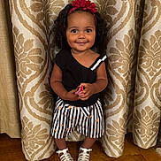 Julia is registered to the contest to win money with this photo: toddler, child, smile, black_top, striped_shorts, heart_pattern, headband, red_flower, curtain, patterned_curtain, white_sandals, wooden_floor, standing, indoors, happy, cute, person, portrait, fashion, toy