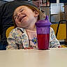 toddler, child, smiling, happy, sippy_cup, purple, red_liquid, floral_jacket, table, indoor, person, seated, yellow_chair, blurred_background, cafe, casual_clothing, small_hands, face, joy, cute
