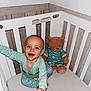 baby, crib, teddy_bear, pajamas, smile, happy, child, indoor, furniture, toy, cushion, person, wood, playful, infant, cute, sleepwear, childrens_room, reaching, portrait