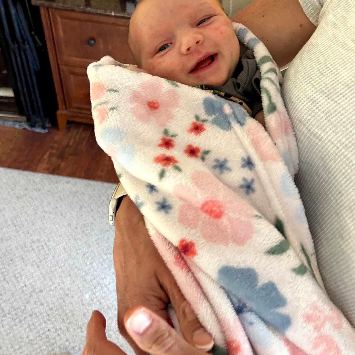 Lorene Hazel joined the competition — help win amazing prizes! baby, newborn, blanket, floral_pattern, smiling, person, adult_hand, holding, indoor, carpet, wooden_furniture, cabinet, white_shirt, skin, face, portrait, cozy, comfort, tenderness, infant