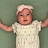 Evie is registered to the contest to win money with this photo: baby, infant, headband, pink, white_dress, floral_pattern, arms_outstretched, green_background, lying_down, cute, face, portrait, child, young, soft_lighting, indoors, skin, person, smile, closeup