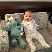 Evie is registered to the contest to win money with this photo: baby, infant, onesie, stuffed_toy, plush, armchair, furniture, cozy, cute, child, indoor, soft_texture, toy, green, white_clothing, portrait, seated, looking_at_camera, home, comfortable