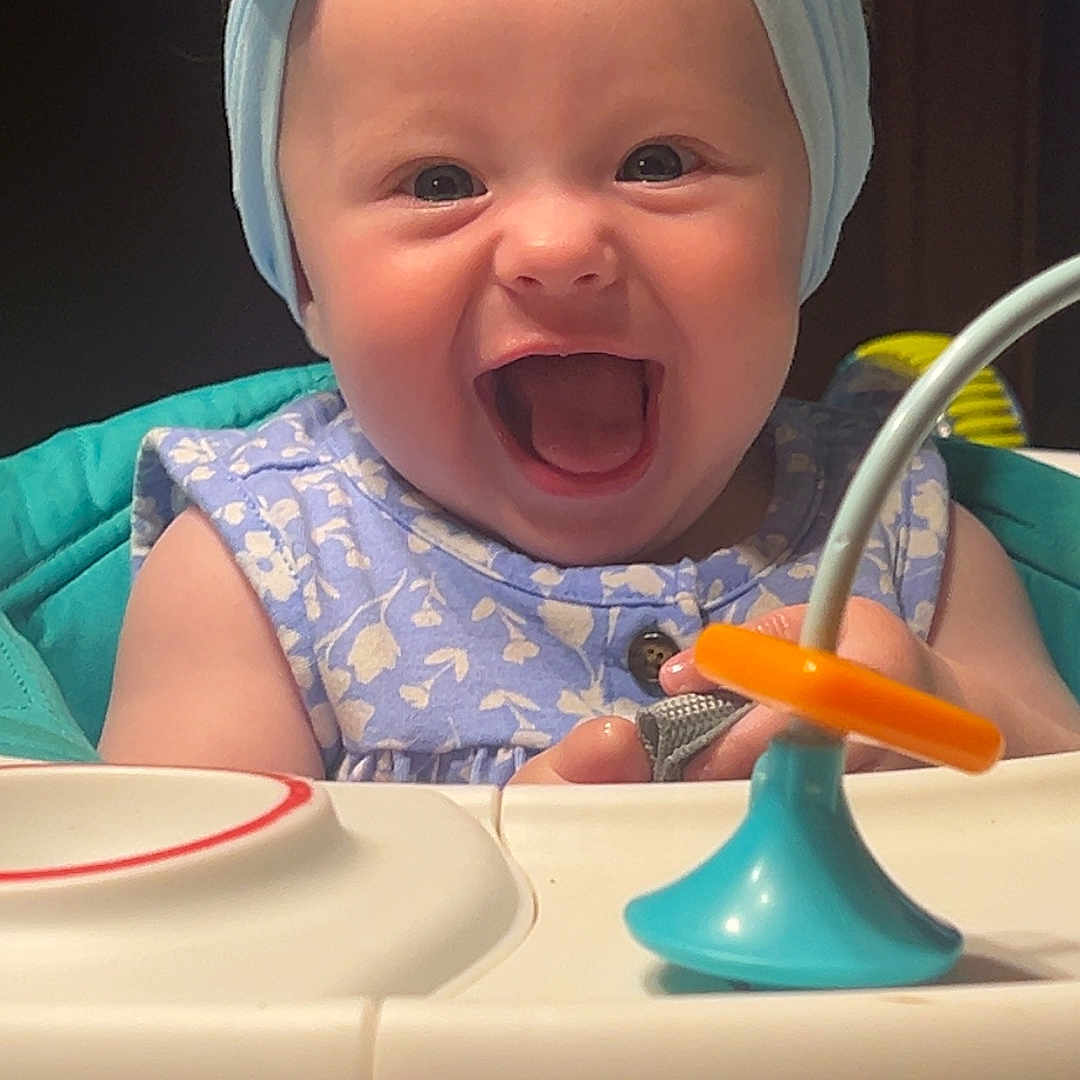 Tilly is registered to the contest to win money with this photo: baby, child, high_chair, headband, blue, toy, smiling, happy, infant, clothing, face, person, cute, indoors, playful, excited, young, seat, expression, joy