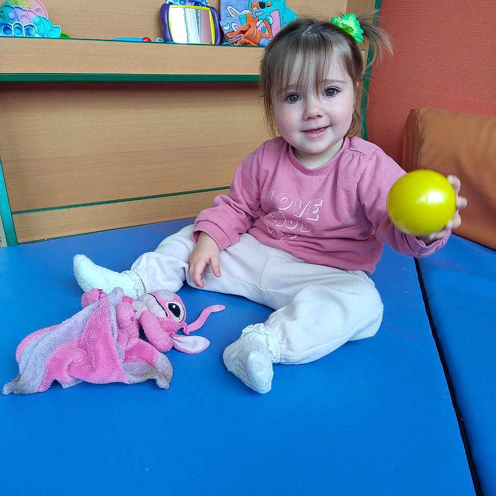Marianna participe au concours pour gagner de l'argent avec cette photo : toddler, child, smiling, yellow_ball, pink_sweatshirt, green_hair_tie, plush_toy, blue_mat, indoor, toys, wooden_shelf, casual_clothing, happy, playtime, person, furniture, cute, young_child, sitting, bright_colors