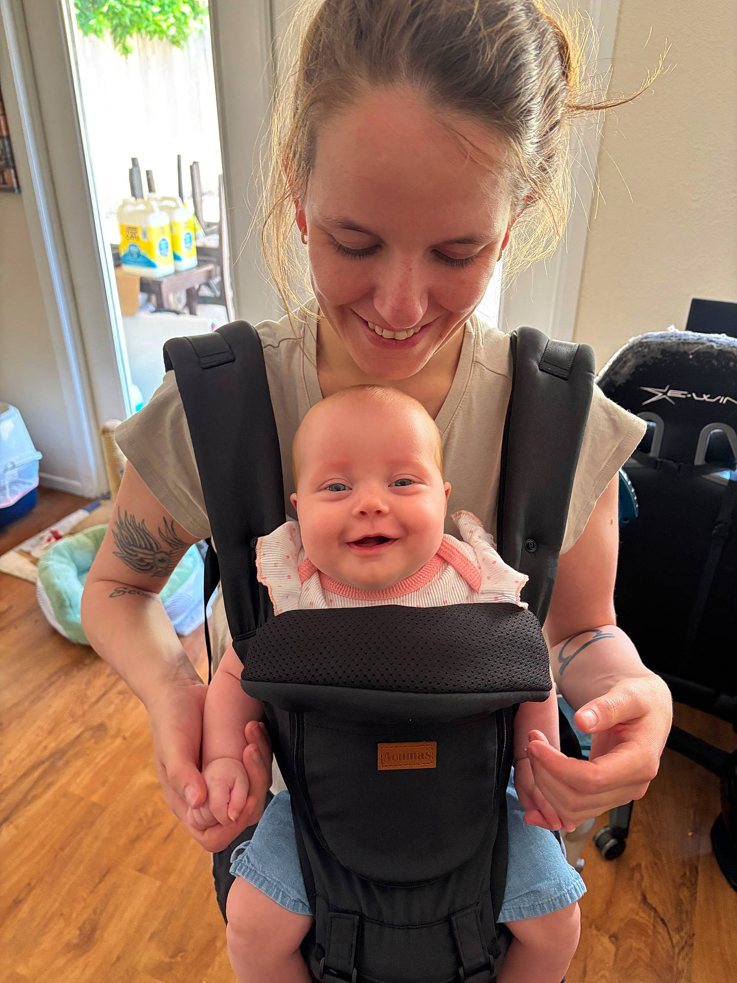 baby, baby_carrier, carrier, casual_clothing, child, face, happy, holding_hands, home, indoor, infant, joy, light, parent, person, shorts, smile, tattoo, woman, wooden_floor