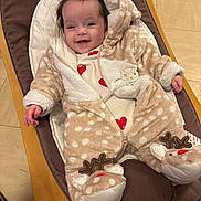 Lila participe au concours pour gagner de l'argent avec cette photo : baby, smiling, fuzzy_onesie, reindeer, indoor, cushioned_chair, wooden_frame, floor_tiles, happy, child, cute, warm_clothing, infant, face, person, cozy, footwear, heart_pattern, hood, relaxed