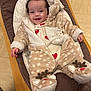 baby, smiling, fuzzy_onesie, reindeer, indoor, cushioned_chair, wooden_frame, floor_tiles, happy, child, cute, warm_clothing, infant, face, person, cozy, footwear, heart_pattern, hood, relaxed