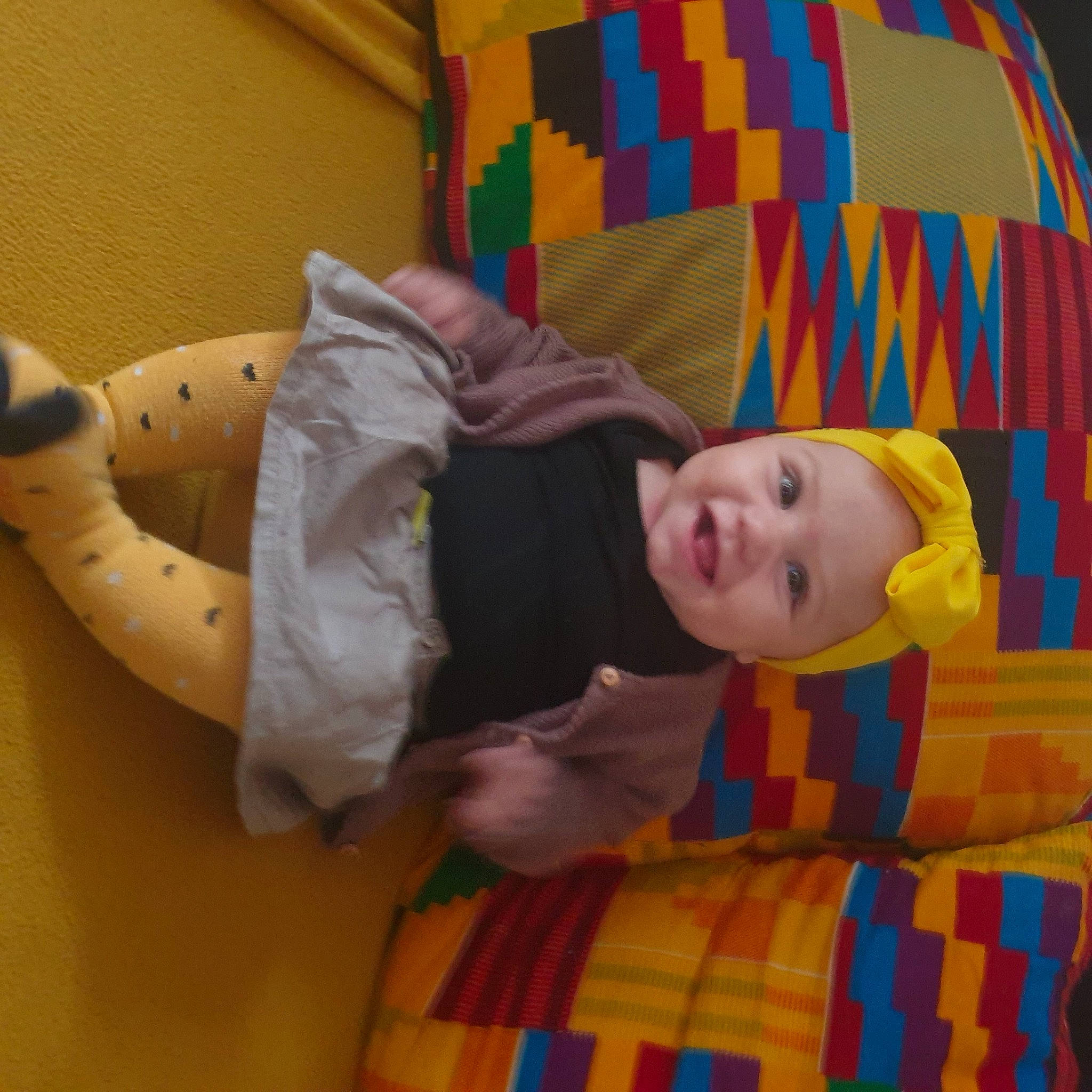 Maelys a rejoint le concours — aidez-le/la à gagner de superbes lots ! baby, baby_toddler_clothing, child, comfort, facial_expression, flooring, fun, headwear, linens, orange, pattern, person, plaid, room, sitting, skin, sleeve, smile, tartan, textile