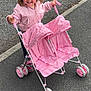 casual_clothing, child, curly_hair, cute, daylight, double_stroller, footwear, fun, hair_tie, happy, outdoor, pavement, person, pink_jacket, play, sidewalk, small_wheels, smiling, toddler, toy_stroller