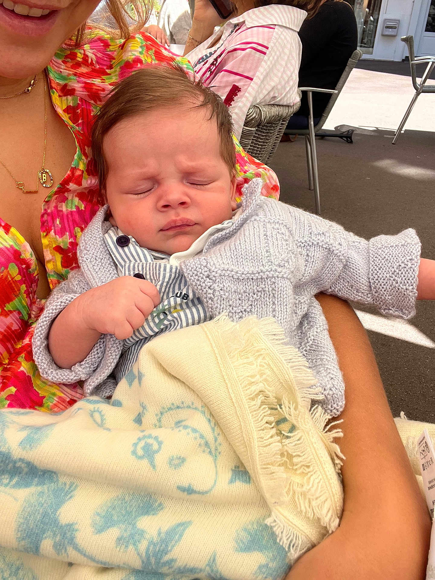 Basile participe au concours pour gagner de l'argent avec cette photo : baby, sleeping, blanket, knitted_sweater, arms, person, floral_clothing, outdoor, cafe, table, chair, sunlight, peaceful, closeup, infant, holding, seated, smiling_adult, casual, relaxed