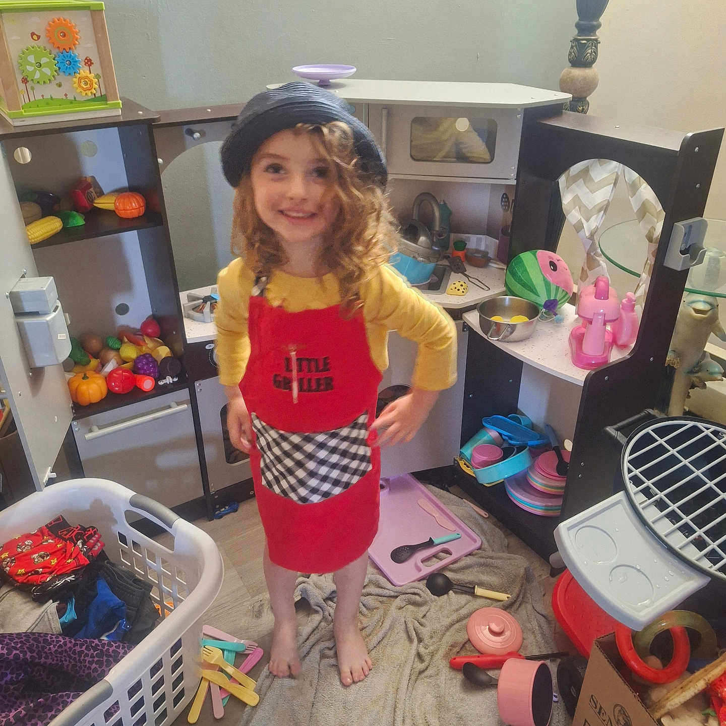 Savannah is registered to the contest to win money with this photo: apron, barefoot, cardboard_box, child, curly_hair, floor, furniture, girl, hat, indoor, laundry_basket, pink_pot, plastic_utensils, playroom, pretend_play, smiling, towel, toy_food, toy_kitchen, yellow_shirt