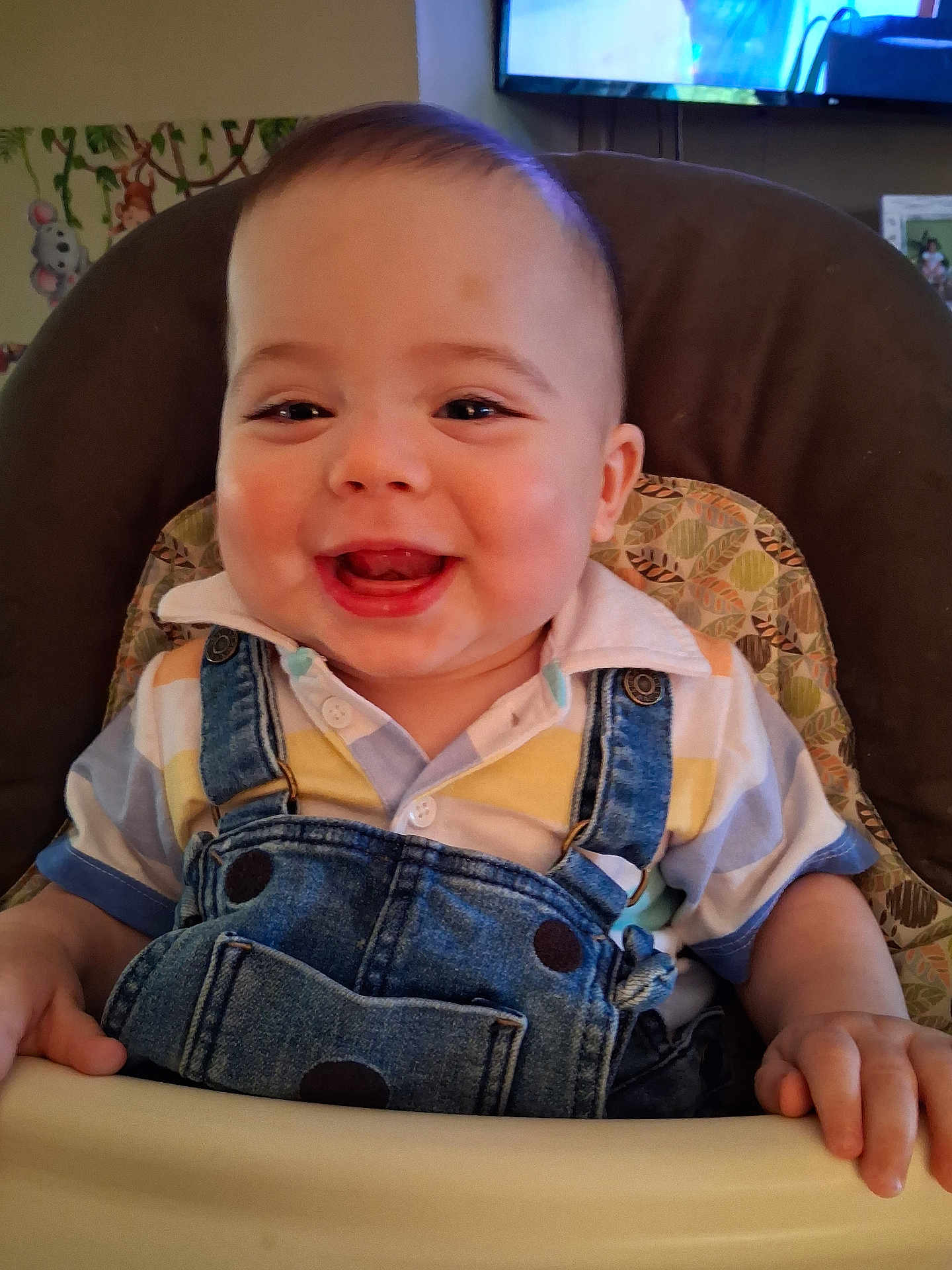 Julius is registered to the contest to win money with this photo: baby, smiling, denim_overalls, striped_shirt, high_chair, happy, indoor, child, face, hands, furniture, television, wall_decor, person, infant, cute, portrait, clothing, home, seat