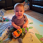 Julius joined the competition — help win amazing prizes! baby, child, toy, stuffed_animal, truck, play_mat, living_room, blanket, carpet, socks, smiling, seated, cute, indoor, furniture, plush_toy, happy, person, young_child, casual_clothing