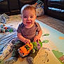 baby, child, toy, stuffed_animal, truck, play_mat, living_room, blanket, carpet, socks, smiling, seated, cute, indoor, furniture, plush_toy, happy, person, young_child, casual_clothing