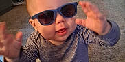 Julius is registered to the contest to win money with this photo: baby, child, sunglasses, striped_clothing, carpet, indoor, living_room, furniture, cabinet, table, chair, person, cute, smiling, hand, legs, floor, home, toy, playful