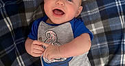 Jaxson is registered to the contest to win money with this photo: baby, blanket, blue, child, clothing, cute, face, gray, hand, happy, head, indoor, infant, lying_down, onesie, plaid, playful, portrait, skin, smiling