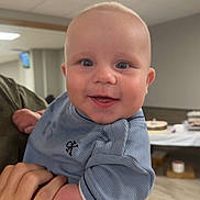 Michael joined the competition — help win amazing prizes! baby, child, infant, smiling, portrait, indoor, held, blue_clothing, onesie, face, eyes, happy, bald, person, adult_hand, ceiling_light, table, party, wood_floor, photography