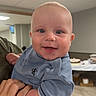 baby, child, infant, smiling, portrait, indoor, held, blue_clothing, onesie, face, eyes, happy, bald, person, adult_hand, ceiling_light, table, party, wood_floor, photography