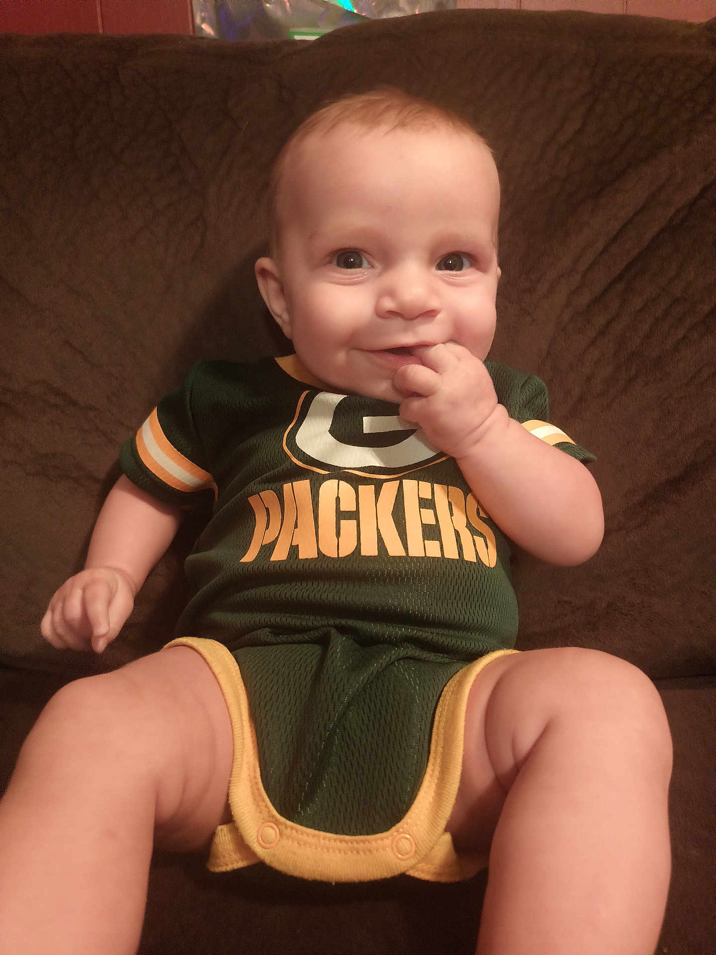 Lainee is registered to the contest to win money with this photo: baby, infant, child, smiling, sitting, green_bay_packers, onesie, clothing, couch, furniture, hand, face, eyes, skin, person, indoors, cute, happy, portrait, baby_fashion