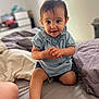 toddler, child, baby, smiling, clapping, bed, blue_clothing, shorts, indoor, person, happy, cute, young_child, curly_hair, skin, face, hands, legs, room, cozy