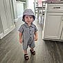 toddler, child, indoor, flooring, clothing, hat, footwear, sandals, cabinet, kitchen, furniture, person, smile, curious, standing, wood_floor, buttoned_shirt, bucket_hat, shorts, young_child