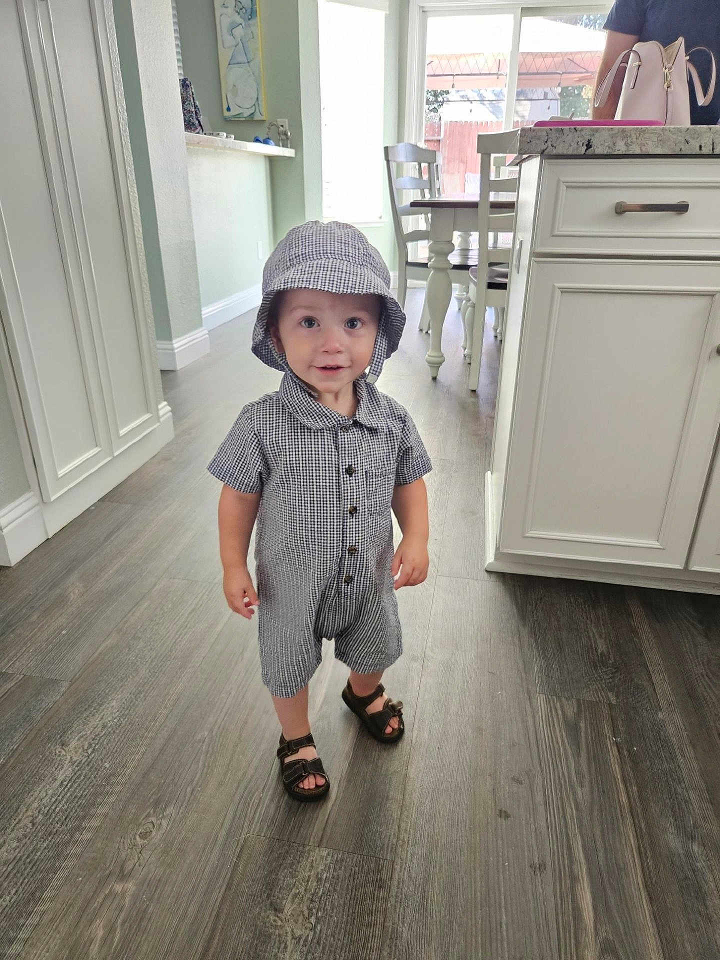 Caden joined the competition — help win amazing prizes! toddler, child, indoor, flooring, clothing, hat, footwear, sandals, cabinet, kitchen, furniture, person, smile, curious, standing, wood_floor, buttoned_shirt, bucket_hat, shorts, young_child