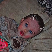 Jerry is registered to the contest to win money with this photo: baby, infant, child, blue_eyes, pillow, blanket, toy, hand, tshirt, football_graphic, lying_down, portrait, smile, cheeks, doodle_marks, fabric, sofa, ear, hair, indoor