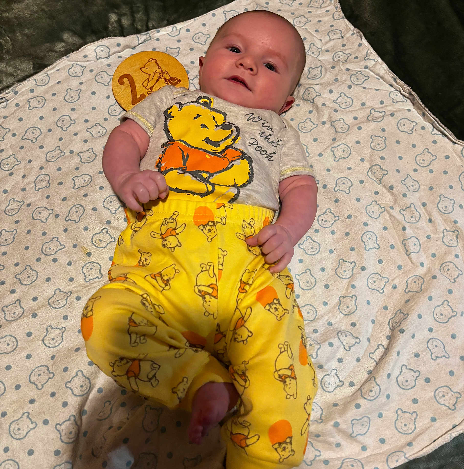 Wyatt is registered to the contest to win money with this photo: baby, infant, winnie_the_pooh, pajamas, blanket, wooden_marker, number_2, clothing, cute, lying_down, face, hand, foot, pattern, yellow, orange, soft_texture, indoors, portrait, child