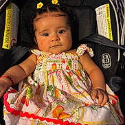 Ivanileth is registered to the contest to win money with this photo: baby, infant, child, portrait, car_seat, floral_dress, yellow_flowers, hair_clips, smiling, sitting, indoors, vehicle, safety_labels, black_background, colorful_clothing, face, eyes, arms, photography, cute