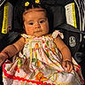 baby, infant, child, portrait, car_seat, floral_dress, yellow_flowers, hair_clips, smiling, sitting, indoors, vehicle, safety_labels, black_background, colorful_clothing, face, eyes, arms, photography, cute