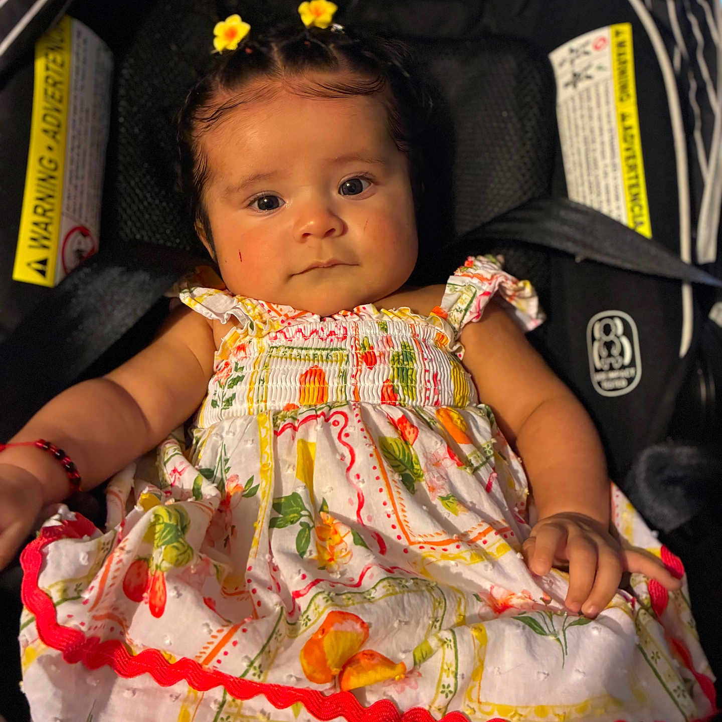 Ivanileth is registered to the contest to win money with this photo: baby, infant, child, portrait, car_seat, floral_dress, yellow_flowers, hair_clips, smiling, sitting, indoors, vehicle, safety_labels, black_background, colorful_clothing, face, eyes, arms, photography, cute