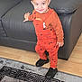 toddler, child, standing, orange_overalls, llama_print, black_sneakers, smile, indoor, couch, leather, carpet, wood_floor, hand, face, hair, clothing, young_child, playful, home, person