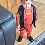 toddler, child, indoor, wooden_floor, orange_clothing, black_vest, sneakers, leather_couch, rug, curious, standing, playful, animal_prints, young_child, home, flooring, casual_clothing, portrait, person, cute