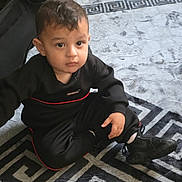Demir is registered to the contest to win money with this photo: toddler, child, black_clothing, sitting, rug, indoor, carpet, footwear, sneakers, curly_hair, serious_expression, floor, young_child, person, home, casual_wear, patterned_rug, baby, cute, portrait