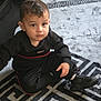 toddler, child, black_clothing, sitting, rug, indoor, carpet, footwear, sneakers, curly_hair, serious_expression, floor, young_child, person, home, casual_wear, patterned_rug, baby, cute, portrait