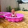 child, toddler, pool, pink_pool, water, summer, outdoor, wooden_deck, plants, fence, sunny, play, happy, splash, curly_hair, swimsuit, young_child, daytime, garden, fun