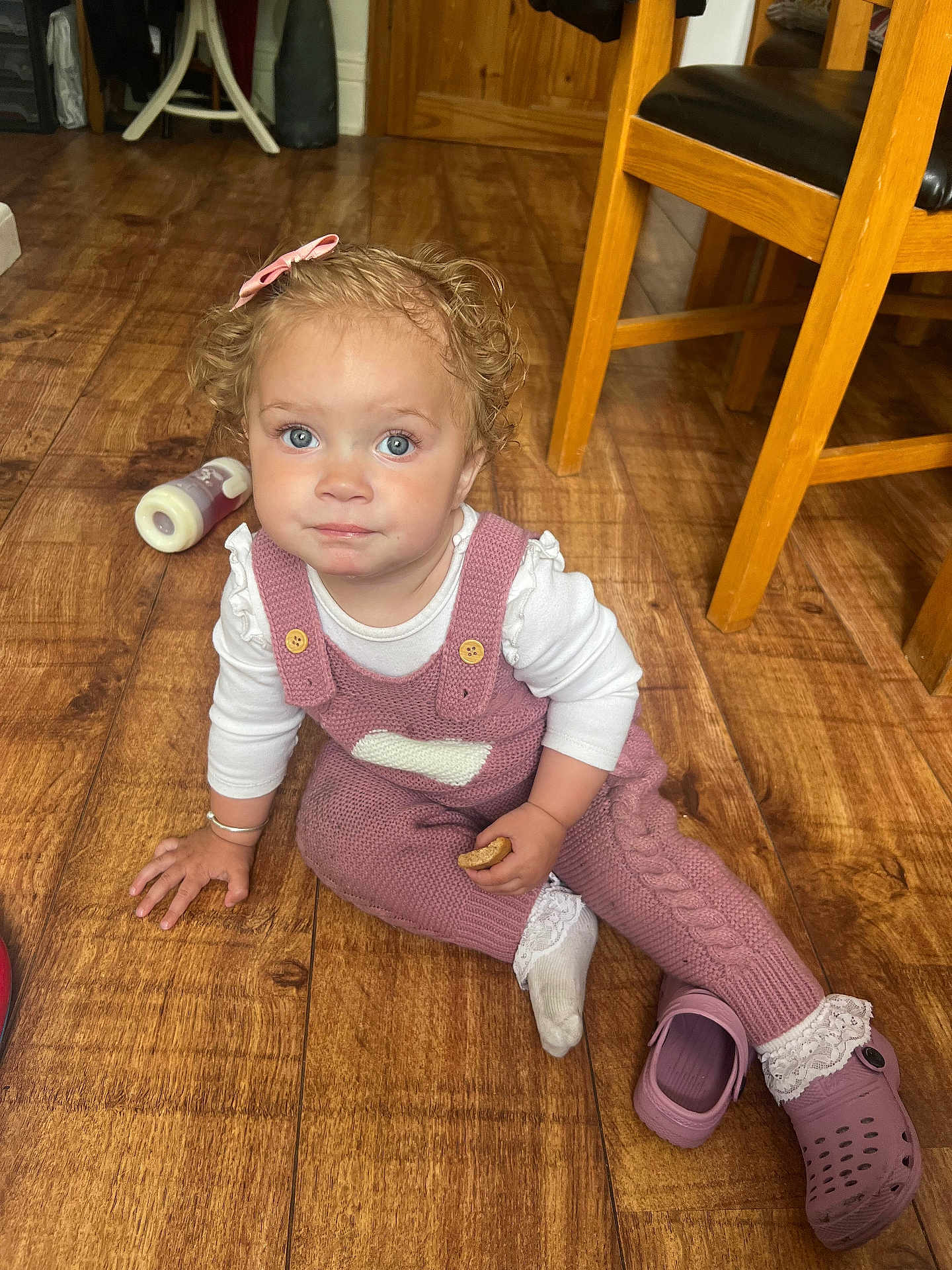 Isabelle is registered to the contest to win money with this photo: toddler, child, baby, curly_hair, blue_eyes, pink_overalls, white_shirt, cookie, wooden_floor, crocs, shoe, indoor, furniture, chair, bottle, flooring, home, cute, portrait, sitting