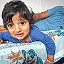 toddler, child, bedspread, floral_pattern, blue_shirt, person, indoor, hand, bracelet, curly_hair, expression, looking, home, furniture, fabric, young_child, portrait, casual, resting, closeup