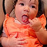 baby, child, tongue, orange_clothing, seat, curly_hair, cute, hand, face, expression, infant, indoor, skin, person, portrait, young_child, sitting, playful, closeup, eyes