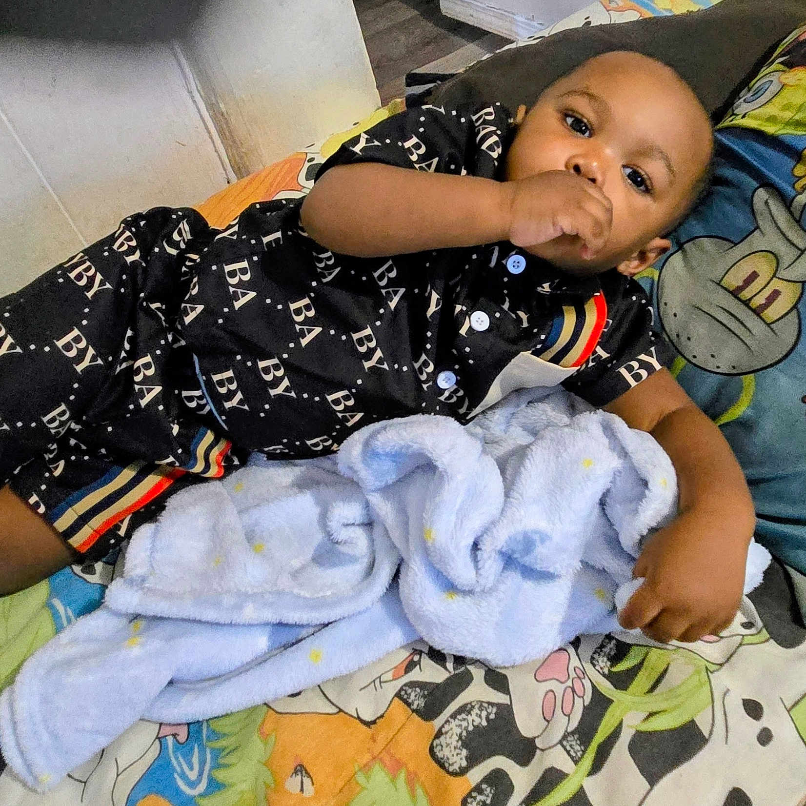 Curtis is registered to the contest to win money with this photo: baby, infant, child, lying_down, portrait, smiling, hand_to_mouth, bed, blanket, pajamas, black_clothing, colorful_bedding, cartoon_pattern, plush_blanket, indoors, home, relaxed, face, single_person, candid_photo